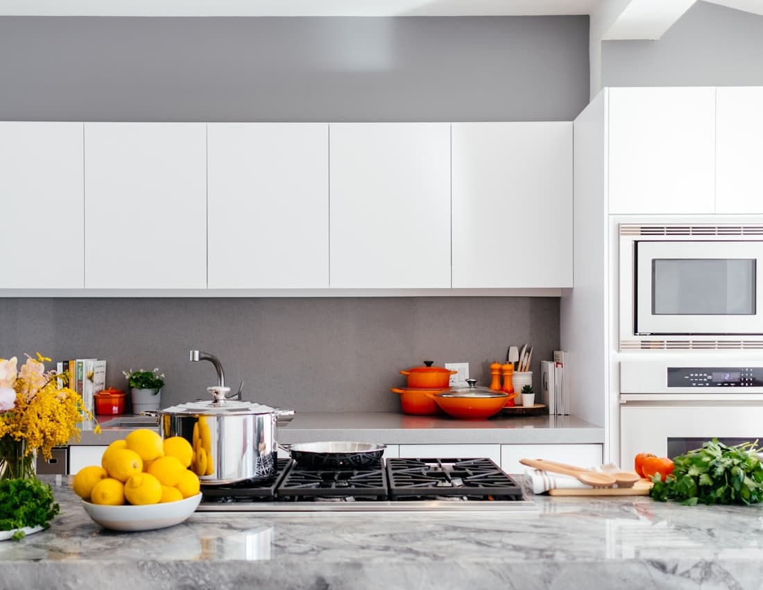 Modern white kitchen with marble countertop, gas stove, orange cookware, and a bowl of lemons in the foreground.
