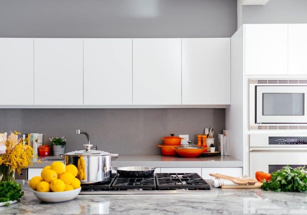 Modern white kitchen with marble countertop, gas stove, orange cookware, and a bowl of lemons in the foreground.