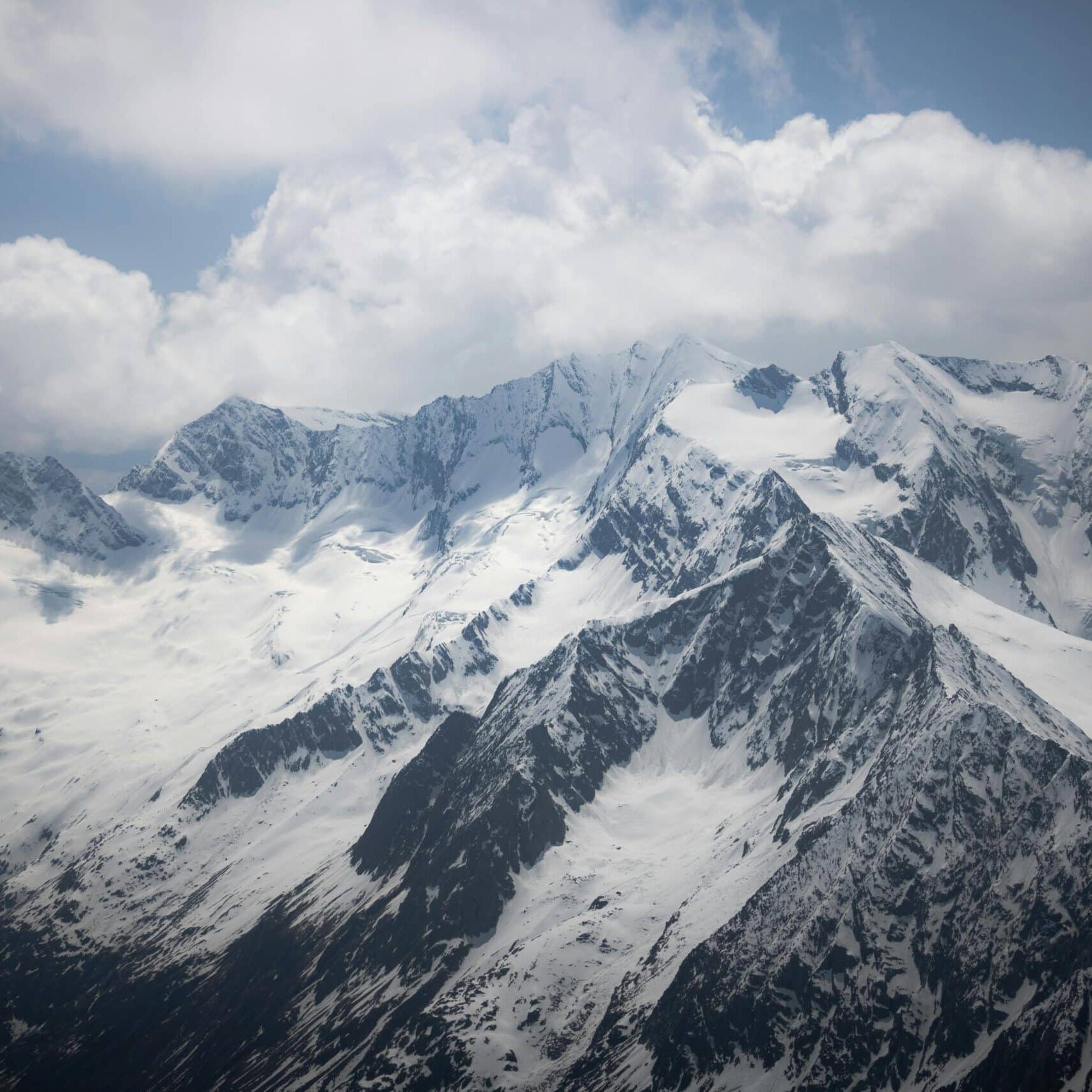 Snow-covered mountain peaks rise dramatically against a cloudy sky, with steep rocky faces and glaciated slopes visible throughout the alpine landscape.